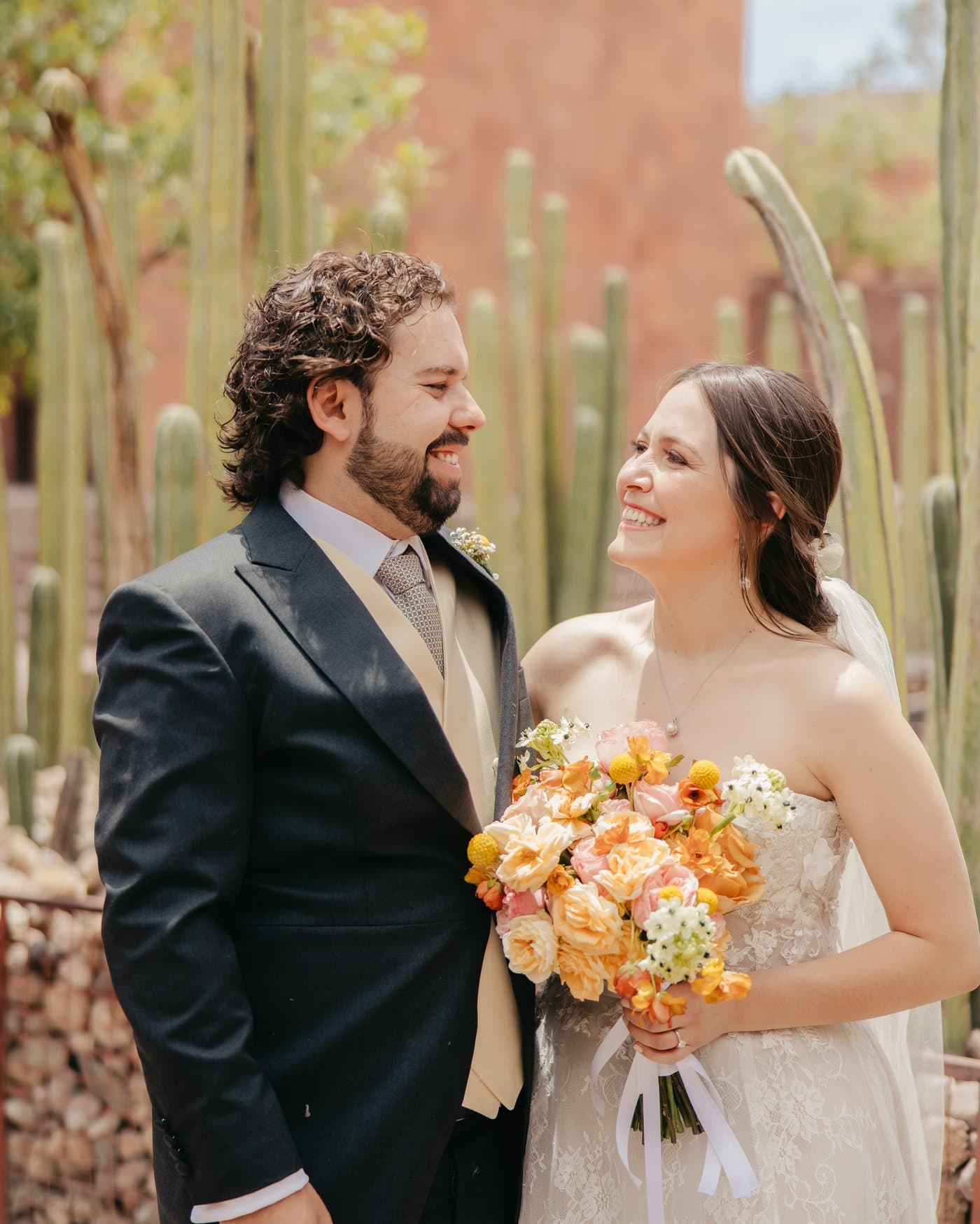 A couple laughing together in front of a terracotta wall and a garden of tall cacti.