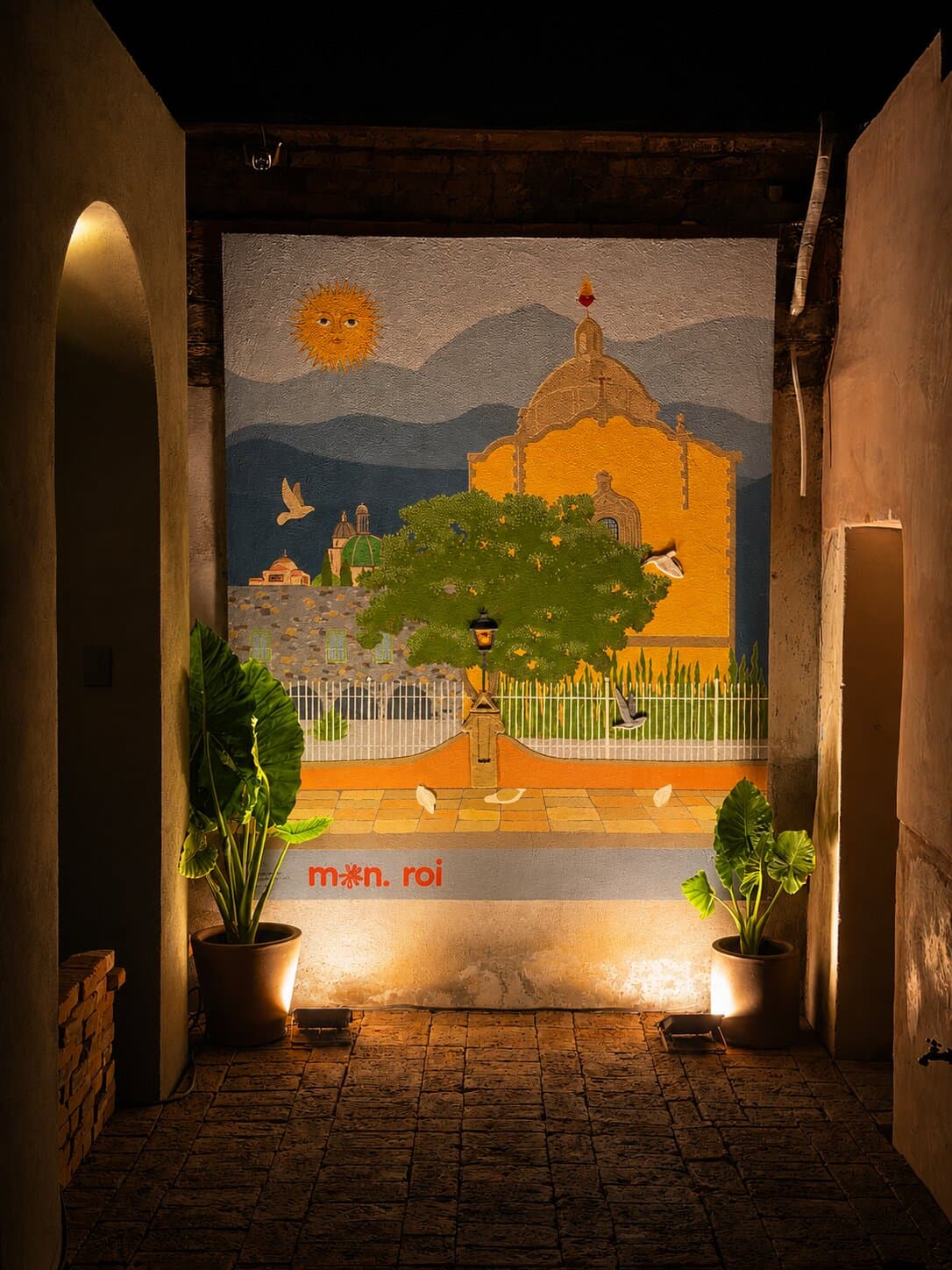 Mural iluminado al final del Pasaje Creativo de Melchor: una cúpula, un árbol y un sol sobre un fondo de montañas, enmarcado por el arco del pasaje.