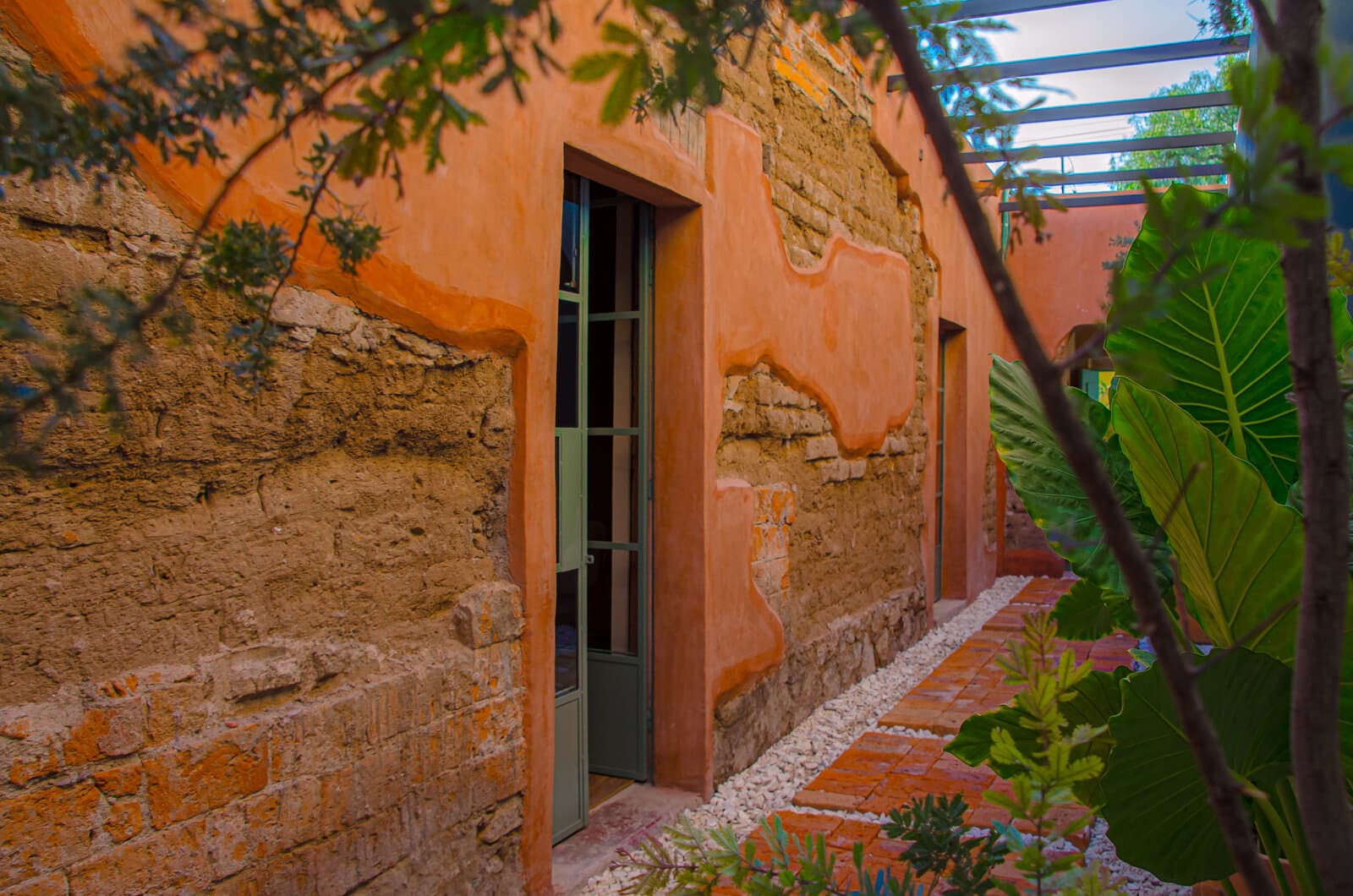 Pasaje exterior de Melchor: muro de adobe y ladrillo bajo acabado terracota, puerta de carpintería verde y camino de ladrillo entre vegetación.