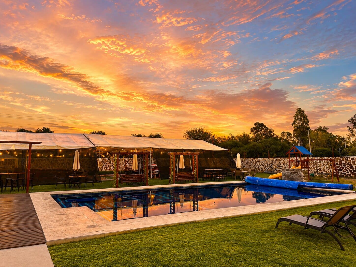 Alberca de Villa Alvento al atardecer, con la terraza iluminada, los columpios y el muro de piedra al fondo.