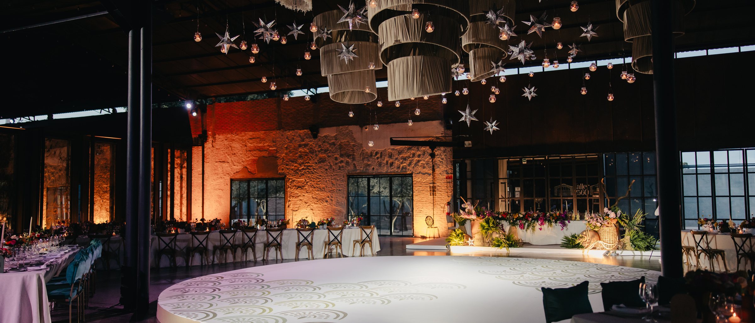 A brick-walled industrial hall at night, lit amber, with woven pendant lights suspended above a circular dance floor.