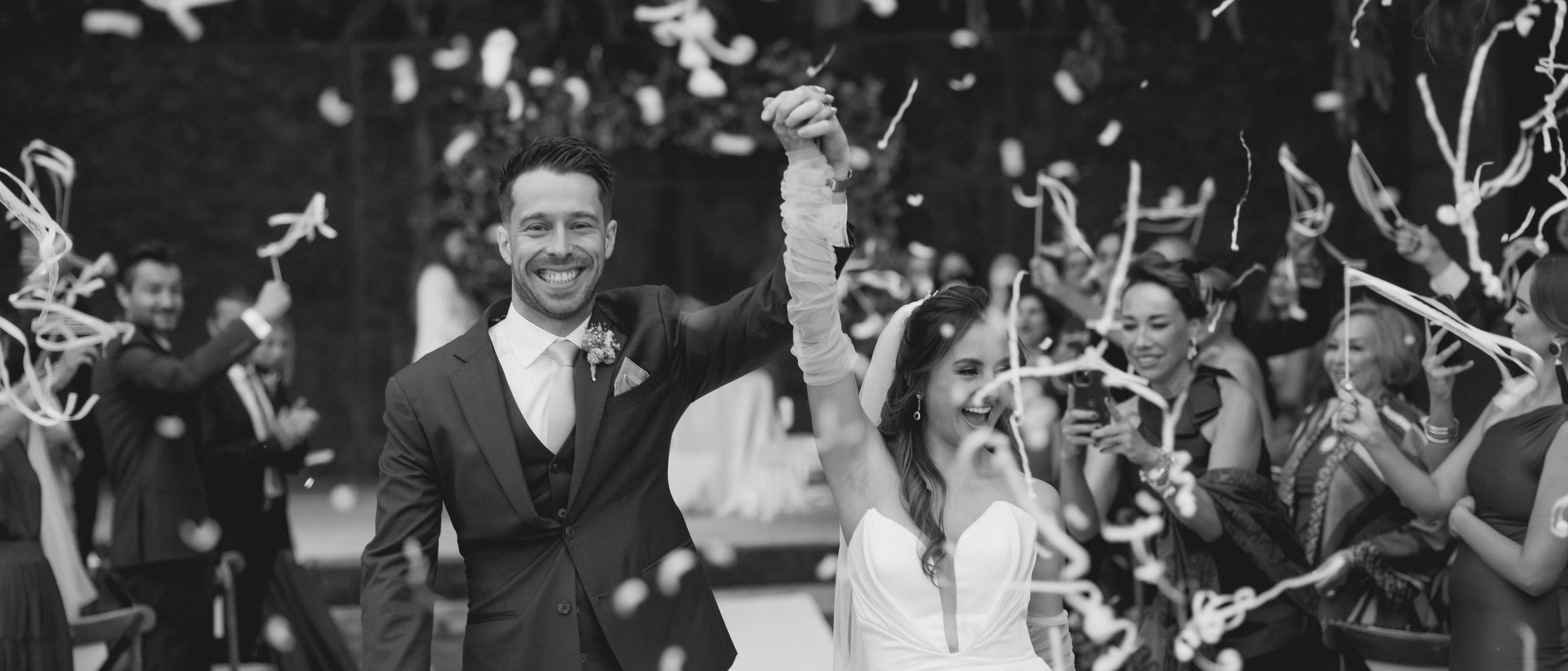 A couple walks hand in hand through a shower of confetti as guests cheer around them, in black and white.