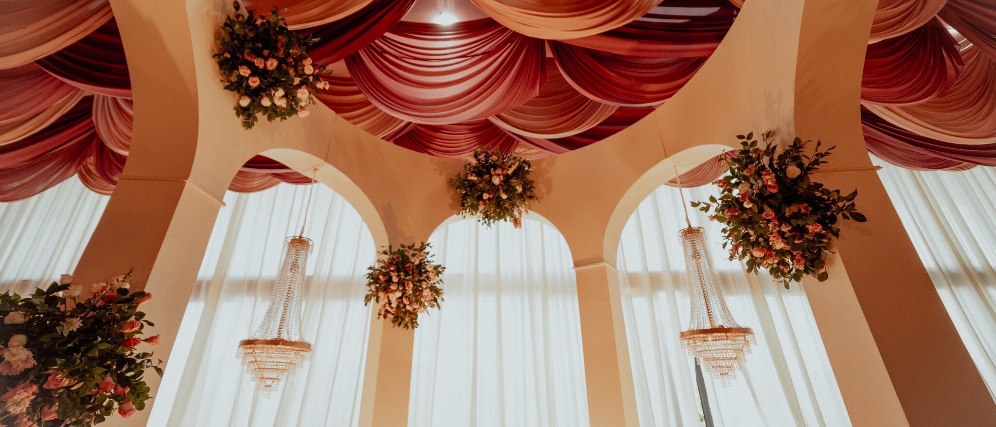 Sculpted columns rising into a suspended fabric ceiling in layered terracotta tones, with crystal chandeliers and hanging floral spheres between them.